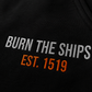 Close-up detail of Burn The Ships EST. 1519 embroidery stitched in white and orange thread on heavyweight black fleece sweatpants.
