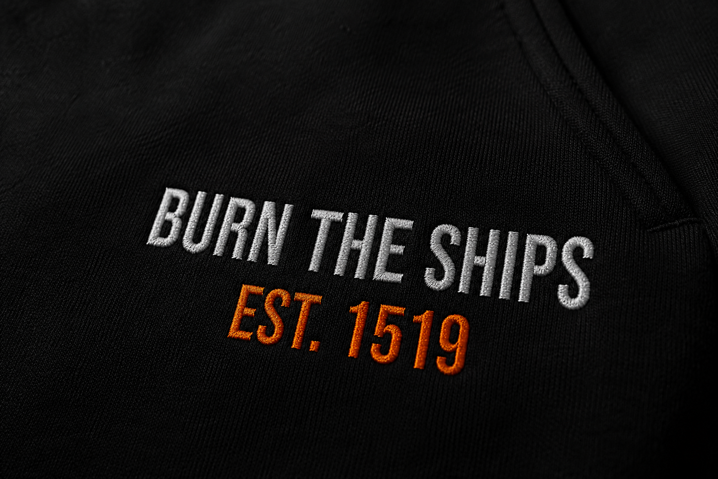 Close-up detail of Burn The Ships EST. 1519 embroidery stitched in white and orange thread on heavyweight black fleece sweatpants.