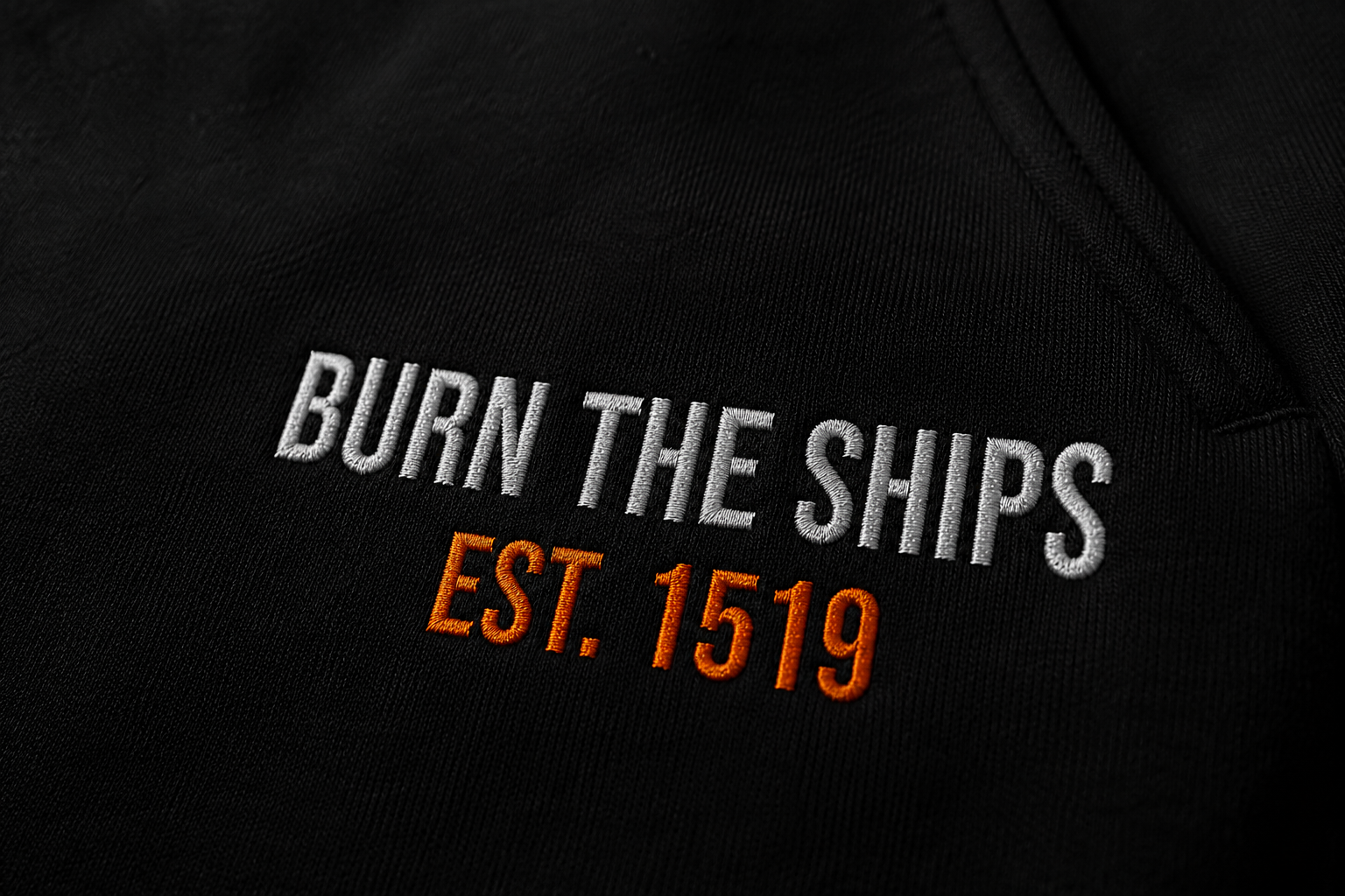 Close-up detail of Burn The Ships EST. 1519 embroidery stitched in white and orange thread on heavyweight black fleece sweatpants.
