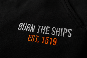 Close-up detail of Burn The Ships EST. 1519 embroidery stitched in white and orange thread on heavyweight black fleece sweatpants.