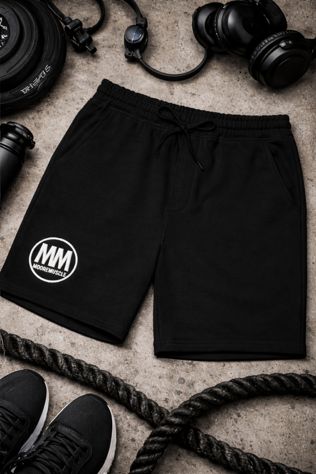 MooreMuscle black fleece shorts laid flat with gym equipment, showcasing embroidered MM logo
