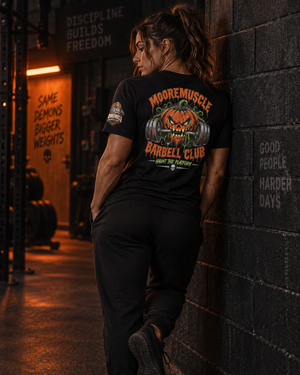 Woman wearing the MMBC Halloween Tee 2026 leaning against a gym wall, MooreMuscle Barbell Club Halloween back print