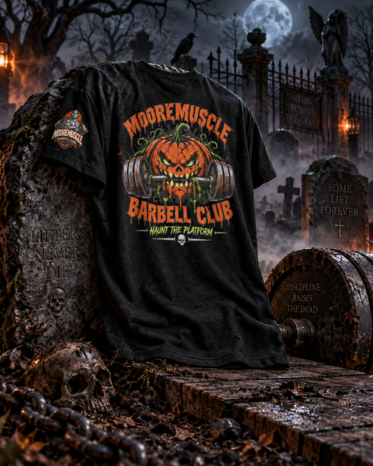 MMBC Halloween Tee 2026 laid flat on a graveyard tombstone, showing the Haunt the Platform pumpkin skull barbell design on a black tee