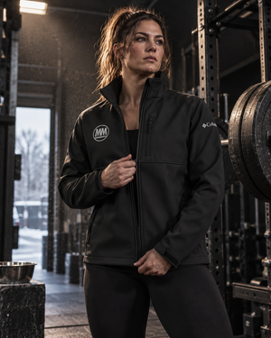 Woman wearing a black MooreMuscle Columbia soft shell jacket inside a powerlifting gym beside weight plates