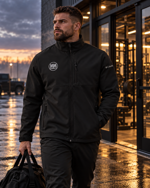 Man in a black MooreMuscle Columbia soft shell jacket carrying a gym bag on a wet city sidewalk at dusk