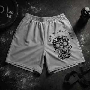 MooreMuscle Day of the Deadlift performance shorts with skull graphic displayed on a gym floor with barbell plate, lifting straps, chalk dust, and shaker bottle