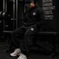 Person sitting on a weight bench in a gym wearing black attire with a MM logo, surrounded by gym equipment.