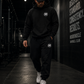 Person wearing a black hoodie and pants with a MM logo, walking in a dark indoor setting.
