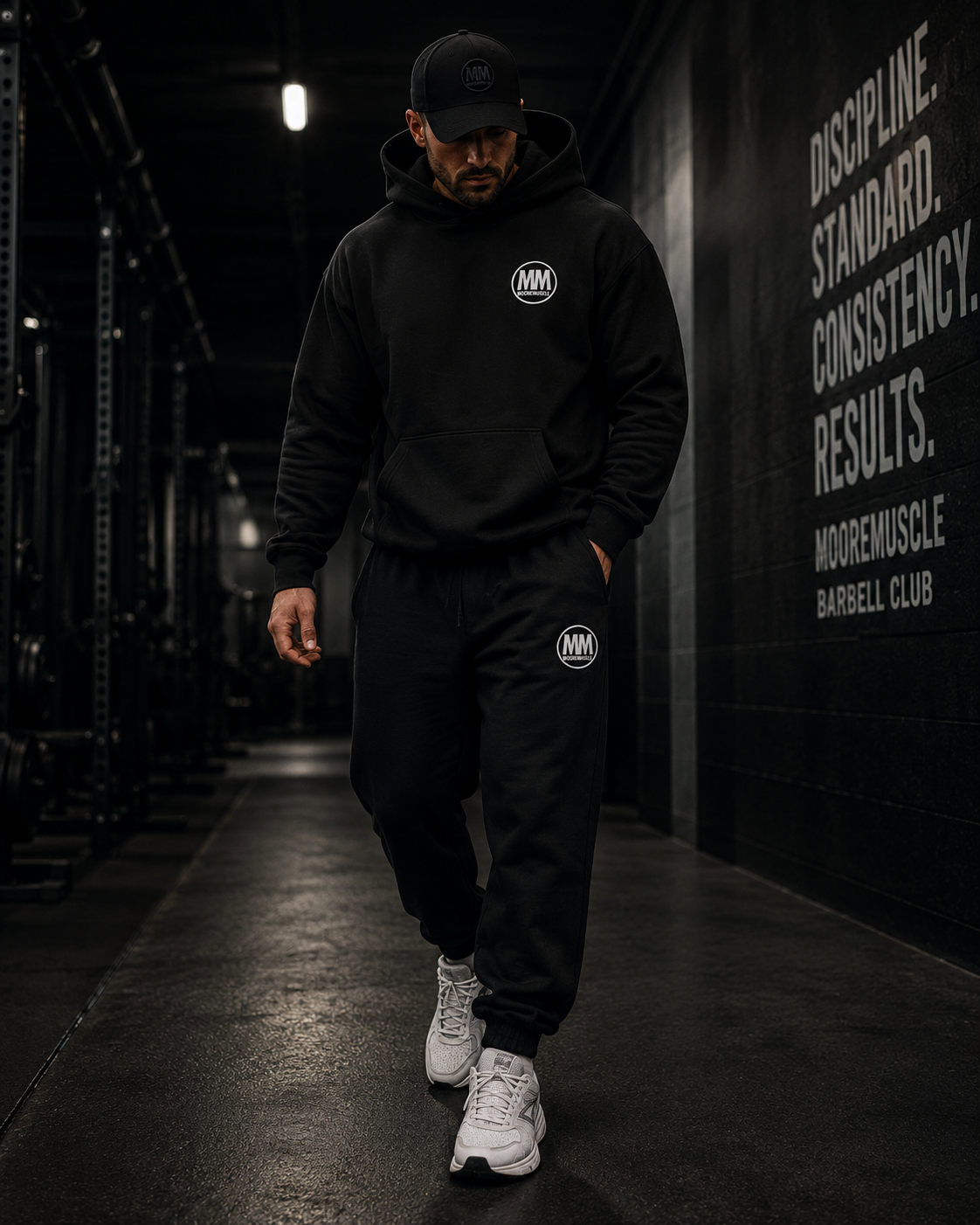 Person wearing a black hoodie and pants with a MM logo, walking in a dark indoor setting.