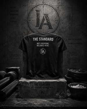 Iron Architect The Standard Tee displayed on a stone pedestal surrounded by architectural details, steel textures, and gym equipment.