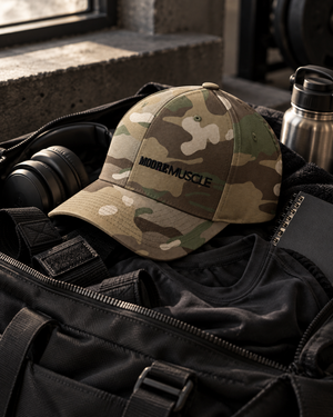 MooreMuscle Uniform Cap in camouflage with black MOOREMUSCLE embroidery, resting on a gym bag beside lifting gear