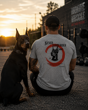 Athlete seated beside a Doberman at sunset wearing the Never Surrender athletic gray tee with the back graphic visible.