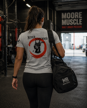Female athlete walking into a strength gym carrying a MooreMuscle gym bag while wearing the Never Surrender athletic gray tee.