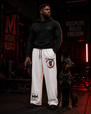 Man in black hoodie and white Never Surrender pants with a dog in a gym setting