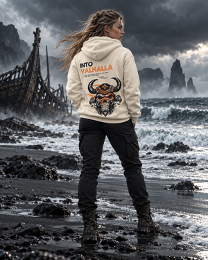 Woman on a stormy black sand beach wearing the Into Valhalla bone hoodie, back view of the horned Viking skull graphic