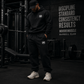 Athlete wearing black MooreMuscle MM Heavyweight Sweatpants and hoodie inside a dark cinematic powerlifting gym with moody lighting and strength culture atmosphere.