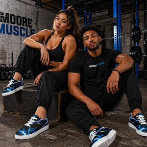 Asian female and Black male athletes wearing MooreMuscle blue lightning low top sneakers in an industrial gym setting, showcasing the flat sole design and bold electric pattern during a strength-focused lifestyle shoot
