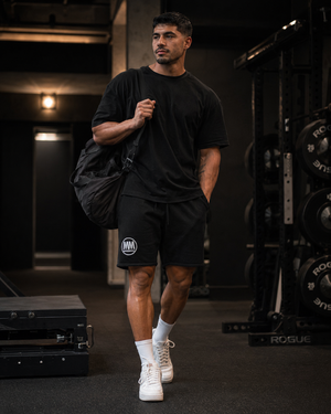Man walking through the gym with a duffel bag, wearing black MooreMuscle fleece shorts with embroidered MM logo
