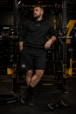 Man wearing black MooreMuscle fleece shorts and matching hoodie with MM logo, leaning against a power rack in the gym