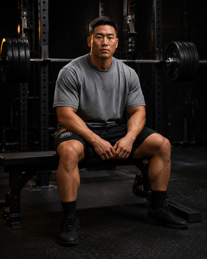 Man seated on a gym bench wearing black MooreMuscle fleece shorts with embroidered MM logo and a gray t-shirt