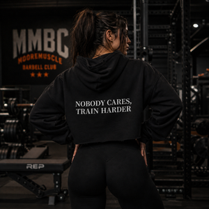 Female athlete wearing black MooreMuscle cropped hoodie back view in gym with “Nobody Cares Train Harder” text