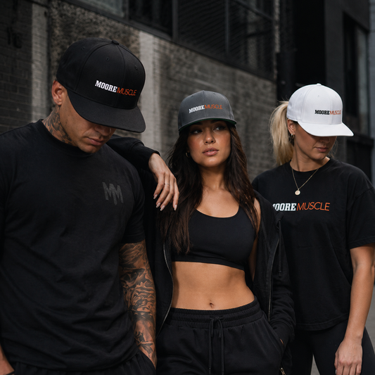 Three models wearing MooreMuscle snapback hats in black, charcoal grey, and white in an urban street setting, styled with athletic apparel against a gritty industrial background