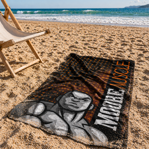 MooreMuscle black and orange stars beach towel laid on sand with natural wrinkles and light sand coverage, ocean waves in the background and a wooden beach chair nearby