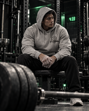 Lifter with his hood up sitting on a bench beside a loaded barbell, wearing the gray MooreMuscle There Is No Spoon hoodie