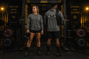 Man and woman wearing MooreMuscle Squat Hooded Long-Sleeve Tees, standing back-to-back in a power rack under Discipline Drives Destiny wall graphics
