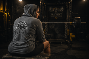 Woman wearing the MooreMuscle Squat Hooded Long-Sleeve Tee, seated back view showing the SQUAT graphic at Priority Health and Fitness