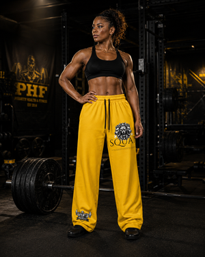 Woman wearing yellow MooreMuscle Squat Sweatpants standing beside a loaded barbell with a Priority Health & Fitness banner in the background